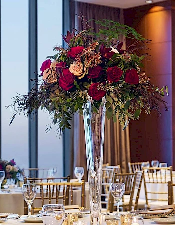 Elegant banquet setup in a high-rise venue with round tables, white linens, glassware, and a tall floral centerpiece bathed in city light.