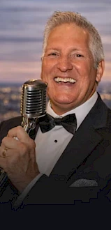 A smiling man in a tuxedo holds a vintage microphone, performing against a city backdrop with a twilight sky.